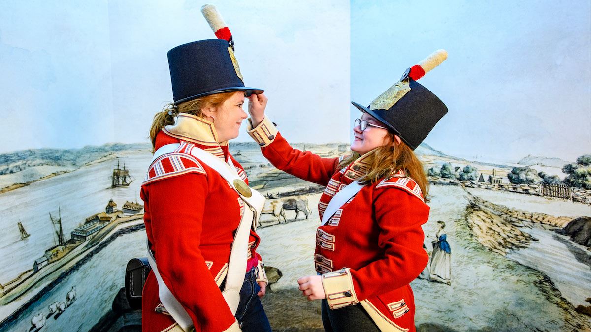A woman and girl facing each other, wearing “red coat” uniforms with tall black “shako” hats. The girl touches the rim of the woman’s hat to straighten it. A painted pioneer-era waterfront scene is in the background.